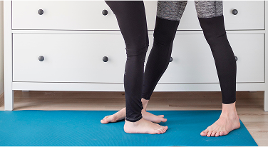 Two people stand barefoot on a teal yoga mat, legs slightly bent, performing a partner yoga pose.