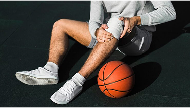 Man sitting on court holding injured knee, basketball nearby