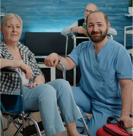 Smiling caregiver assists senior woman in wheelchair, another senior in background.