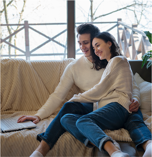 Couple cuddling on a couch, looking at a laptop. Cozy home scene.
