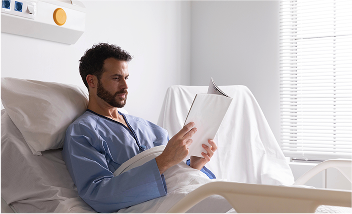 Man in hospital gown reading a magazine in bed.